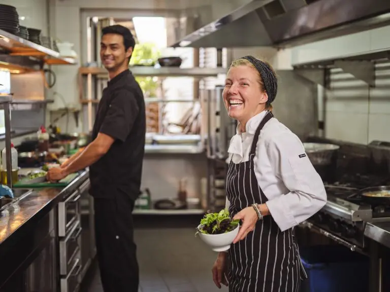 Female chef in a commercial kichen holding a bowl of greens and smiling. A kichenhand is in the background cutting up vegetables.
