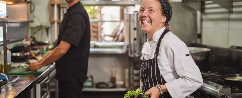 Female chef in a commercial kichen holding a bowl of greens and smiling. A kichenhand is in the background cutting up vegetables.