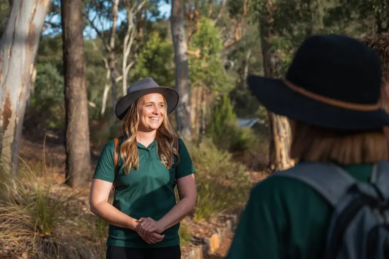 Female tour guide wearing wide-brimmed hat and green polo shirt standing outdoors with gum trees behind her