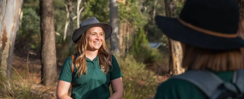 Female tour guide wearing wide-brimmed hat and green polo shirt standing outdoors with gum trees behind her